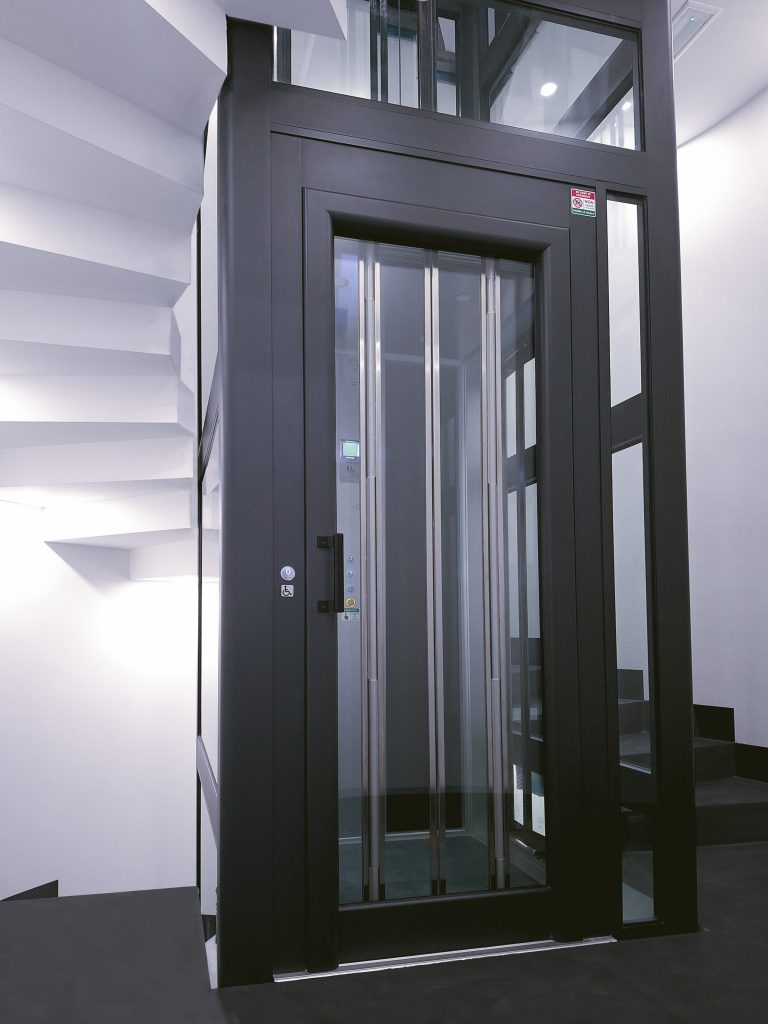 Home lift Eltec Lift HLB elettrico in vano circolare con gradini a sbalzo, vetro trasparente e struttura nera TA ; design elegante e integrazione perfetta tra architettura e funzionalità. Vista a contrasto frontale.