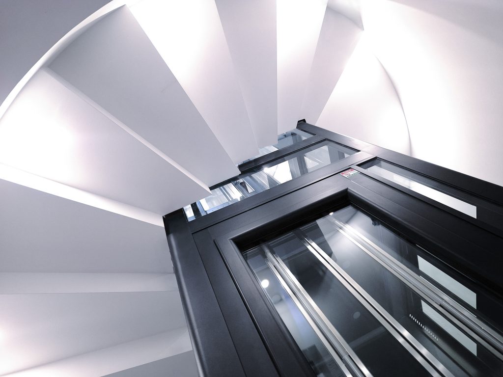 Home lift Eltec Lift HLB elettrico in vano circolare con gradini a sbalzo, vetro trasparente e struttura nera TA ; design elegante e integrazione perfetta tra architettura e funzionalità.