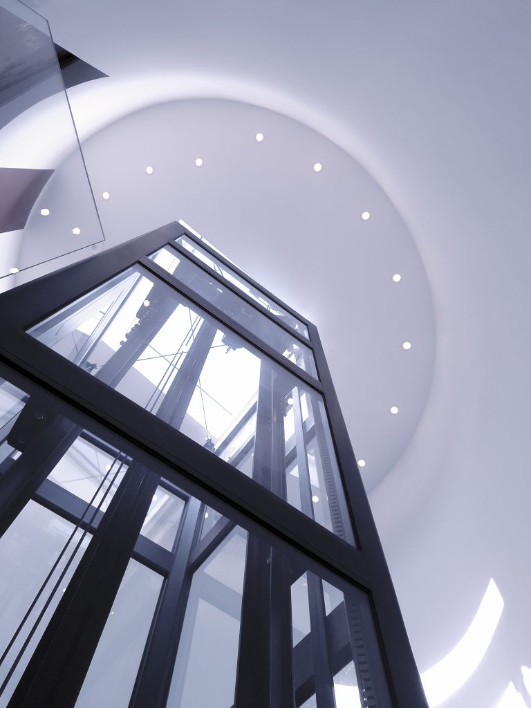 Home lift Eltec Lift HLB elettrico in vano circolare con gradini a sbalzo, vetro trasparente e struttura nera TA ; design elegante e integrazione perfetta tra architettura e funzionalità. Vista verso il soffitto e illuminazione.